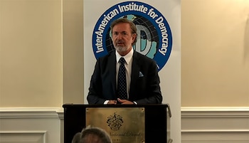 Hombre de traje y corbata con barba canosa habla desde un podio. Detrás, el logo del Instituto Interamericano para la Democracia en un banner