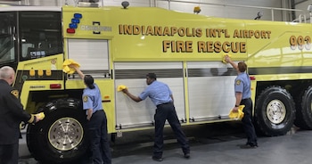Cuatro bomberos limpian el costado de un gran camión de bomberos amarillo con la inscripción "Indianapolis Int'l Airport Fire Rescue" en un hangar
