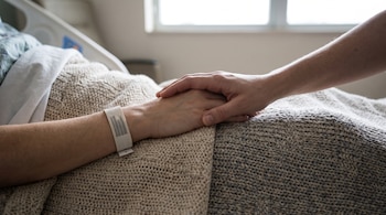 Una mano sostiene a otra que tiene una pulsera médica de identificación, reposando sobre una manta en una cama de hospital.