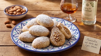 Galletas con azúcar glas en un plato decorado, junto a un tazón con almendras, una copa, una botella y una tarjeta con texto.