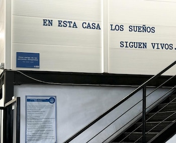 Pared blanca con texto. Cartel azul con logo UNHCR y 'Casa amiga de las personas refugiados'. Cartel 'Reglamento de dormitorio'. Escalera metálica
