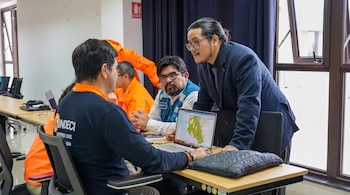 Cuatro hombres en una sala; uno sentado con camisa azul de Indeci usa una laptop que muestra un mapa. Otro hombre de pie observa la pantalla