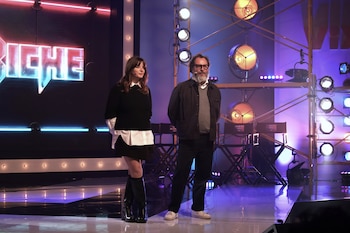 Una mujer y un hombre con barba están de pie en un escenario junto a focos de estudio y sillas de director