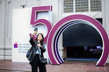 Mujer sonriente tomando una selfie con su smartphone frente a un gran cartel morado que muestra el número '50' y parte del logo de la Feria del Libro