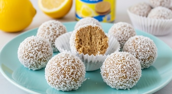 Primer plano de trufas de limón cubiertas de coco rallado en un plato azul claro. Una trufa está abierta en un capacillo. Al fondo se ven limones frescos y una lata.