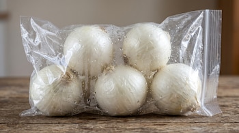Una bolsa de plástico sellada con gotas de agua en su interior contiene cinco cebollas blancas sobre una superficie de madera.