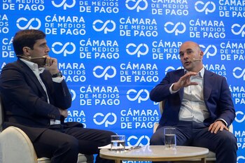 Dos hombres con traje conversan sentados ante un fondo azul con el texto Cámara de Medios de Pago