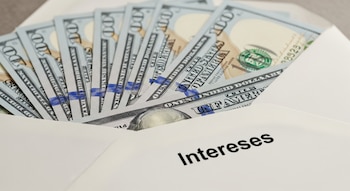 Varios billetes de cien dólares estadounidenses asoman de un sobre blanco sobre una superficie gris. El sobre lleva la palabra 'Intereses' en español.