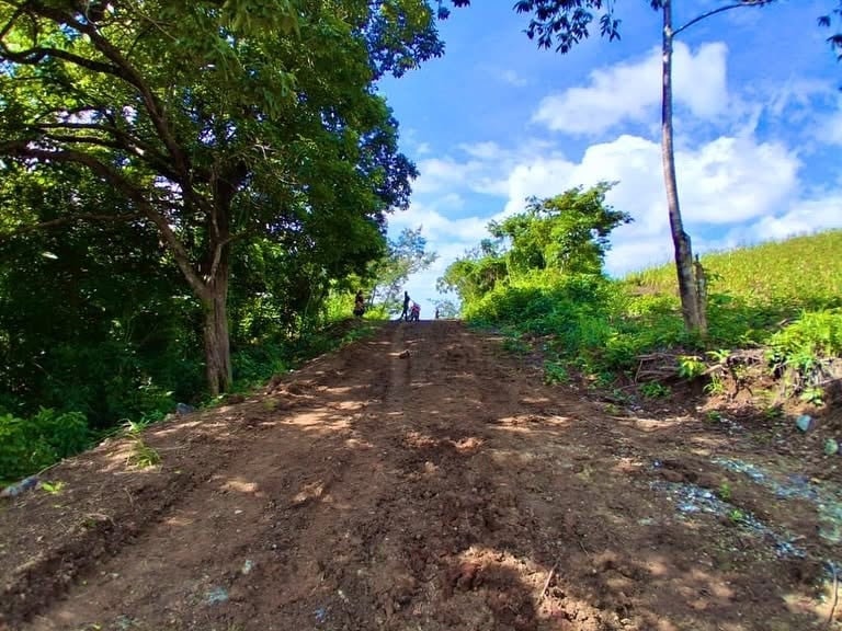Un sendero de tierra recién trabajado asciende por una colina verde rodeado de árboles frondosos y vegetación exuberante bajo un cielo azul vibrante con nubes blancas. (Cuerpo de Ingenieros del Ejército de Guatemala)