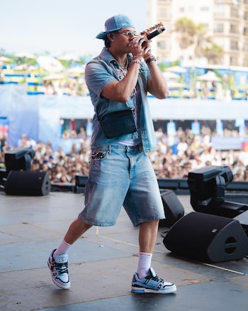 Hombre con gorra, camisa y bermudas de mezclilla, gafas de sol y joyas, sostiene un micrófono en un escenario con altavoces y una multitud al fondo