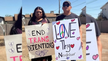 Dos personas, una con cabello largo oscuro y otra con gorra, sostienen carteles de protesta bajo un cielo azul claro, abogando por los derechos trans y la paz