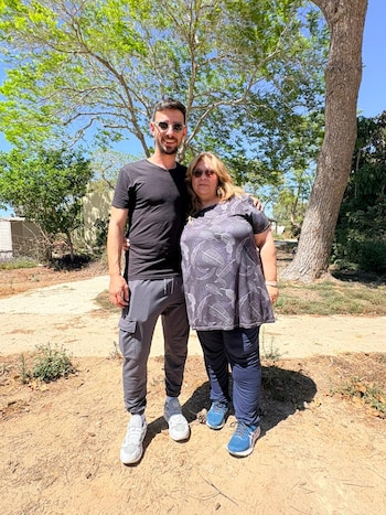 Hombre joven con barba y mujer de mediana edad con cabello rubio posan abrazados al aire libre bajo un cielo azul claro, rodeados de árboles y tierra
