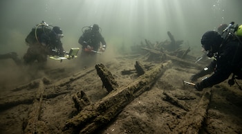 Tres buzos provistos de equipos de inmersión e instrumentos de medición inspeccionan vigas de madera sumergidas en el lecho marino.