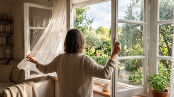 Una persona de espaldas abre una ventana blanca con marco de madera con vistas a un jardín, junto a una taza y una cortina blanca.
