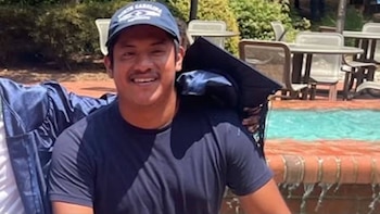 Hombre joven con gorra de béisbol sonríe sosteniendo toga y birrete de graduación. Al fondo hay una piscina y mesas con sillas