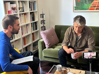 Emmanuel Carrère, de mediana edad y cabello entrecano, sentado en un sofá verde sosteniendo una taza, frente a un entrevistador joven con papeles, siendo grabado por un smartphone
