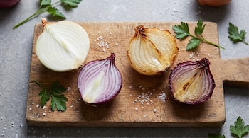 Cuatro mitades de cebolla blanca y morada, crudas y cocinadas, sobre una tabla de madera con sal y perejil.