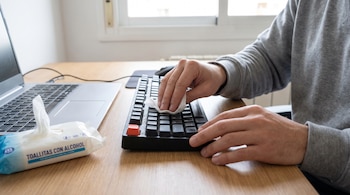 Manos de una persona limpian el teclado negro de una computadora con una toallita blanca. Hay un paquete de toallitas con alcohol y una laptop.