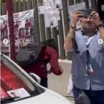 Un hombre con chaleco graba con un teléfono frente a una reja con carteles de personas desaparecidas y un automóvil en primer plano
