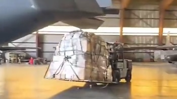 Montacargas transporta pila grande de paquetes envueltos bajo el ala de un avión de carga en un hangar. Se observa otra aeronave al fondo