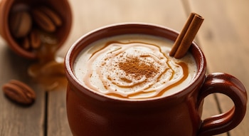 Primer plano de atole de cajeta en taza de barro con canela en polvo, sirope y dos ramas de canela. Cajeta derramada y nueces en madera.