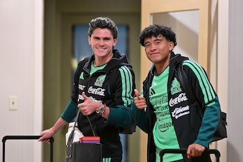 Dos futbolistas con uniforme deportivo verde y negro sonríen, hacen el gesto con el pulgar hacia arriba y sujetan sus maletas