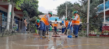 Un grupo de trabajadores con chalecos naranjas, cascos verdes y botas de goma usa escobas para limpiar un patio escolar con agua. Hay un edificio y árboles al fondo