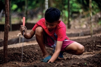 Um menino de cabelos pretos, vestindo camiseta vermelha e shorts rosa, está pulando no chão escuro, usando ferramentas laranja para cultivar.