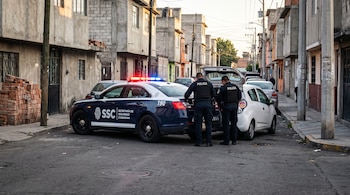 Dos policías de espalda inspeccionan el maletero de un auto blanco junto a una patrulla con torreta encendida en una calle residencial.