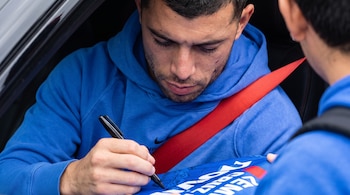 Un hombre con sudadera azul y cinturón de seguridad rojo firma una prenda de tela azul con un marcador negro.