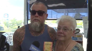 Un hombre barbudo con gafas de sol y camiseta azul sostiene un billete de lotería Powerball ante un micrófono, junto a una mujer de cabello blanco y gafas