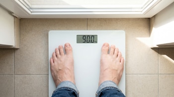 Vista aérea de los pies de una persona descalza, con el dobladillo de unos jeans, de pie sobre una balanza digital blanca que marca 90.0 kg en una pantalla.