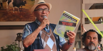 Hombre con sombrero, gafas y camisa a cuadros sostiene micrófono y libro, un hombre con barba a la derecha, pintura de vacas en el fondo