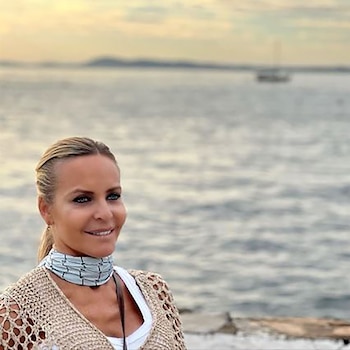 Primer plano de Alejandra Forlán, mujer rubia con el pelo recogido, sonriendo, con un top de ganchillo. Al fondo, el mar y un barco bajo un cielo claro