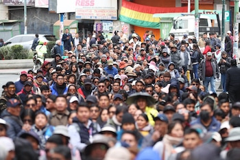 Primer plano de un oficial de la Policía Boliviana con equipo antidisturbios asistiendo a una mujer mayor de cabello gris; otros agentes en línea detrás