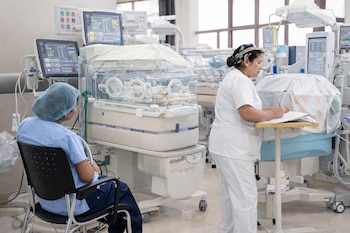 Dos mujeres, una sentada y otra de pie, varias incubadoras con bebés, monitores médicos, equipo hospitalario, silla, mesa y ventanas.