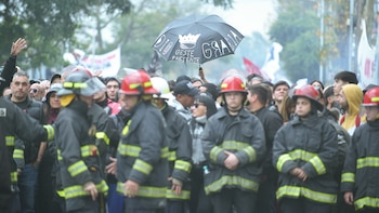 Una multitud de personas, incluyendo varios bomberos con uniforme, se congrega al aire libre; en el fondo, un paraguas negro lleva un diseño blanco