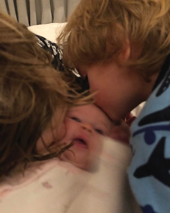 Un niño con cabello rubio besa la frente de un bebé. Otro niño con cabello castaño está parcialmente visible. Los niños están en una cama blanca