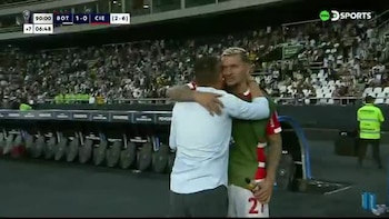 Un jugador de fútbol con camiseta verde y roja abraza a un hombre con camisa clara al borde de la cancha en un estadio