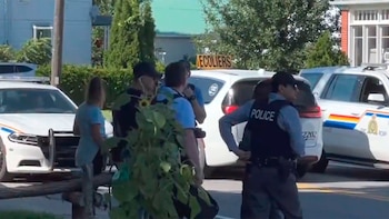 Agentes de policía vestidos con uniformes y chalecos tácticos están de pie junto a dos coches patrulla blancos con las luces apagadas en una calle residencial arbolada