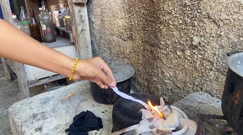 Una mano con pulsera amarilla sostiene una tira blanca sobre un recipiente con fuego, junto a ollas metálicas y estantes con frascos.