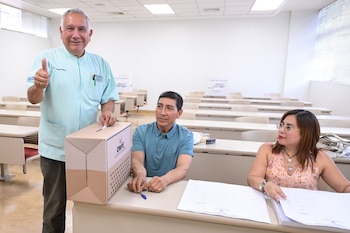 Armando Massé, en camisa celeste, deposita un voto en una urna de ONPE, sonríe y da pulgar arriba. Dos personas están sentadas en la mesa de votación