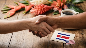 Dos manos se estrechan sobre una mesa de madera rústica; una tarjeta con la bandera de Costa Rica, un adhesivo de Estados Unidos, flores y café caliente.