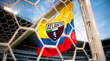 Bandera de Colombia con escudo de Atlante FC en la portería de fútbol. La red está en primer plano, con un estadio y luces de fondo.