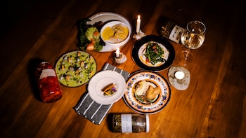 Mesa de madera, siete platos con alimentos, dos velas, copa de vino, frascos de conservas, botella de hierbas, servilleta rayada