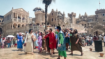 Actores con túnicas interpretan una escena bíblica frente a un castillo de piedra con torres y balcones; espectadores y un cielo azul claro completan la imagen