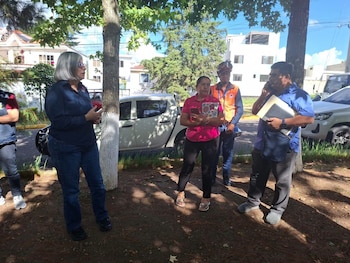 Cinco personas están de pie bajo árboles. Una mujer de cabello gris habla a otras. Hay una camioneta blanca y un SUV blanco estacionados, y edificios residenciales detrás