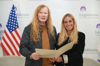 Un hombre con cabello rubio largo y una mujer sonríen a cámara mientras sostienen un documento; la bandera de Estados Unidos se ve detrás