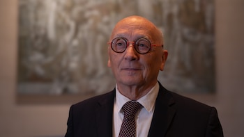 Primer plano de un hombre calvo y canoso de edad avanzada, con gafas de montura redonda roja y negra, vistiendo un traje oscuro y corbata estampada, con un pin negro en la solapa