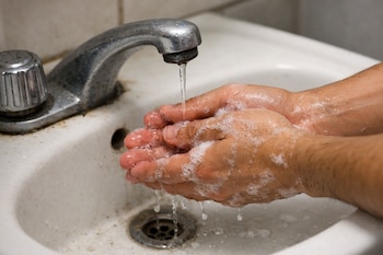 Dos manos con espuma de jabón bajo un grifo metálico que vierte un chorro de agua escaso sobre un lavabo blanco.